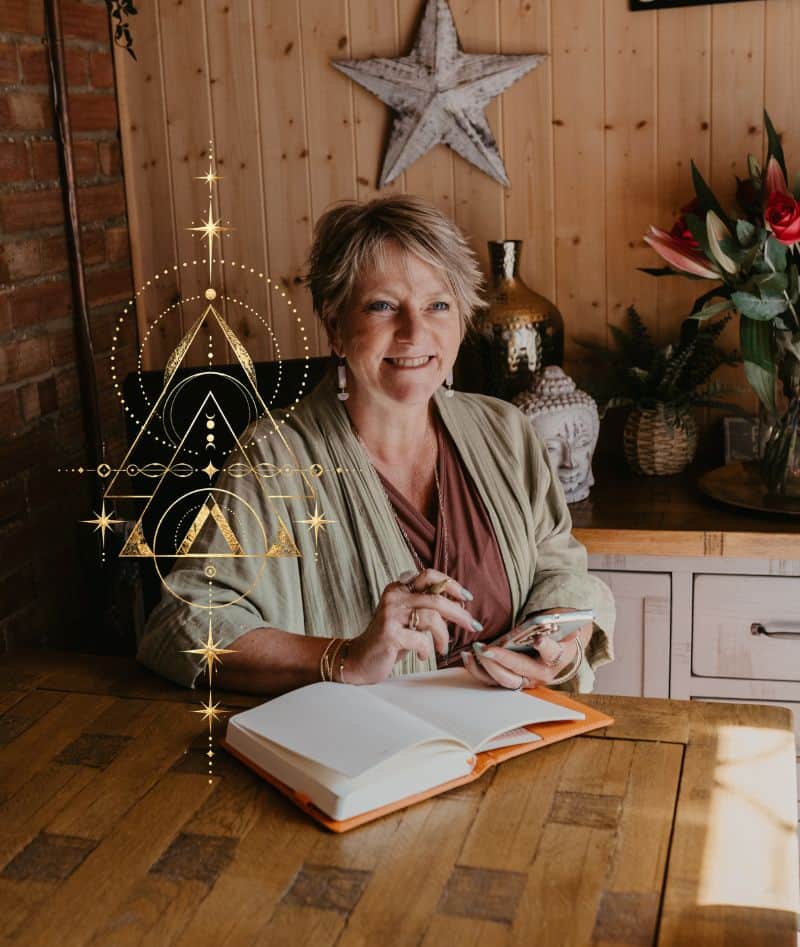 Tania Meacher smiling at a wooden table, holding her phone and a pen beside an open notebook.