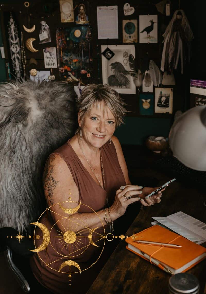 Spiritual WordPress Web Design - Creative Witch Designs (1) Tania Meacher smiling at her desk, holding a phone beside an open notebook in her creative workspace.