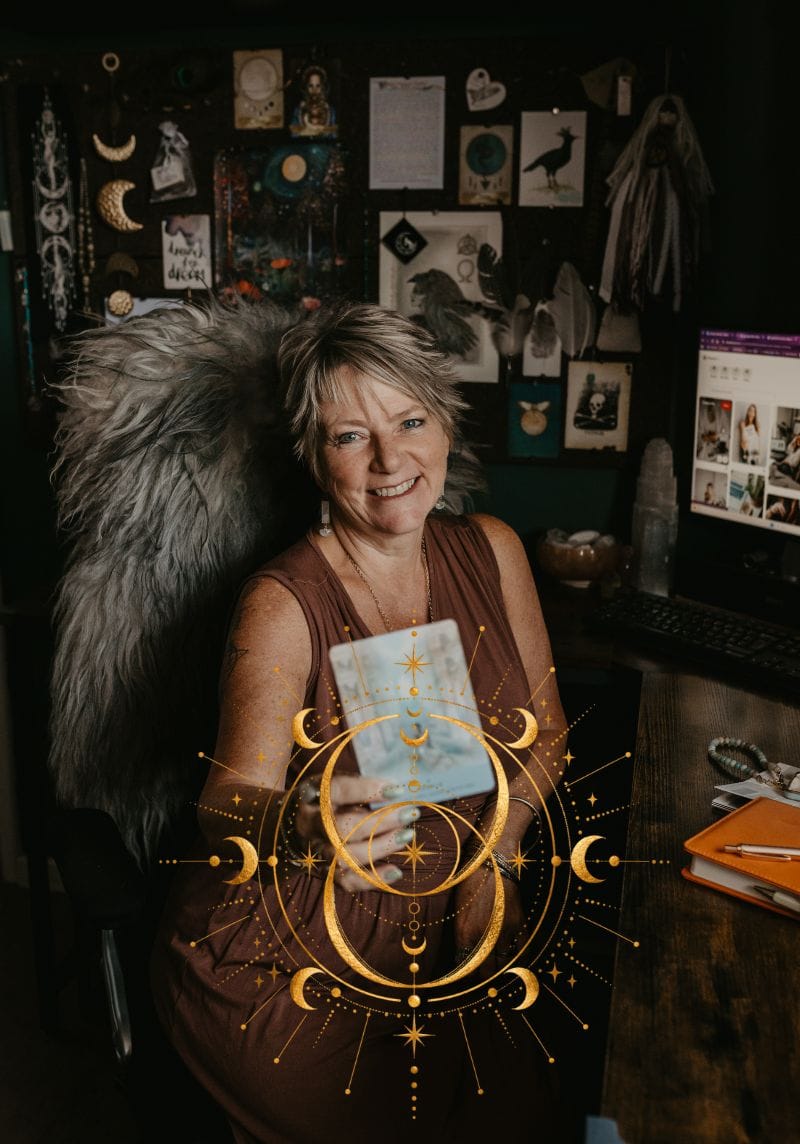Tania Meacher smiling at her desk while holding up an Oracle card, with her computer and notebook beside her.