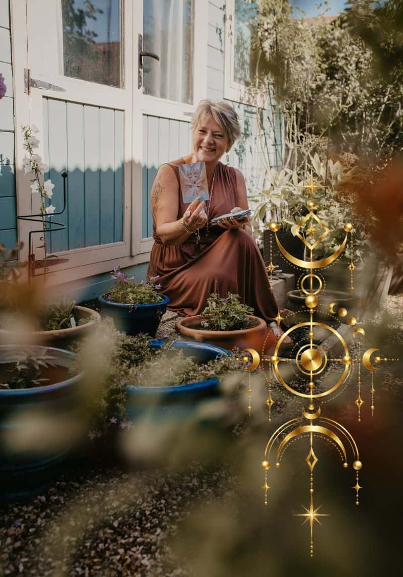 Tania Meacher smiling outside her garden studio, holding a card and a deck of cards among potted plants.